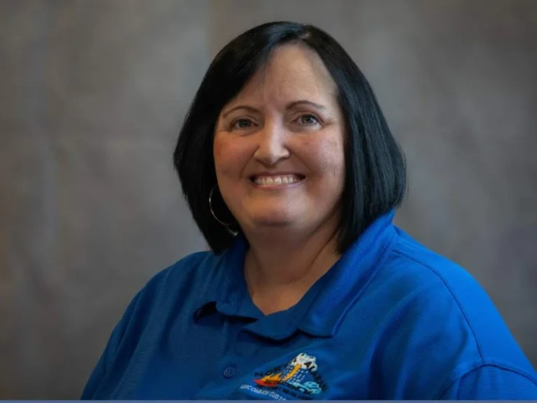 Smiling woman with black hair wearing a blue polo shirt against a gray background.