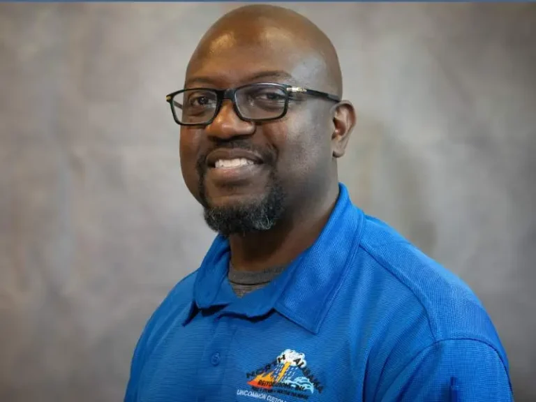Man with glasses smiling, wearing a blue collared shirt against a gray background.