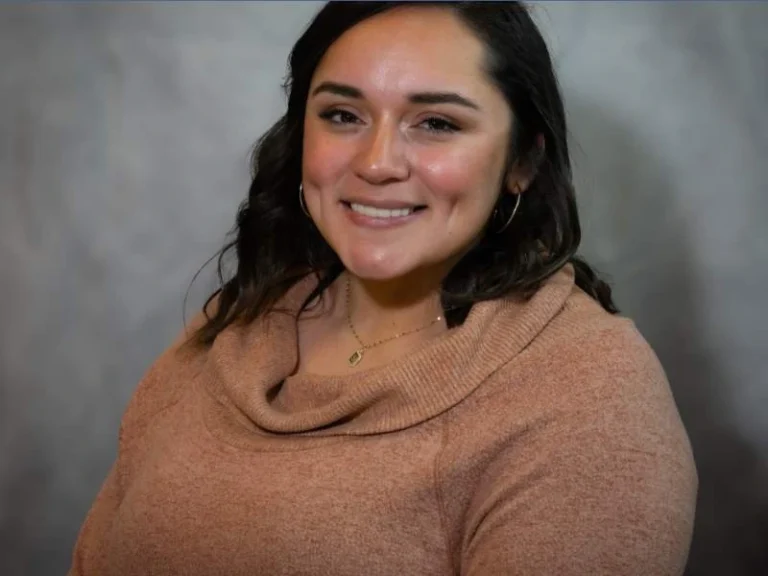 Smiling woman with dark hair wearing a brown sweater and gold hoop earrings against a gray background.
