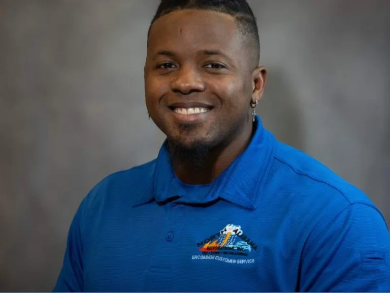 Smiling man wearing a blue polo shirt with a logo and text on the chest against a gray background.