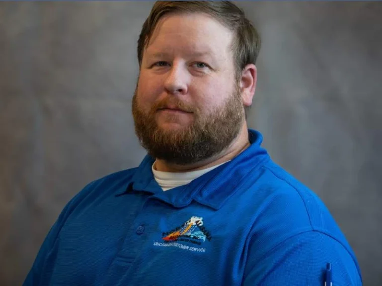 Bearded man wearing a blue polo shirt with a logo, posing against a gray background.