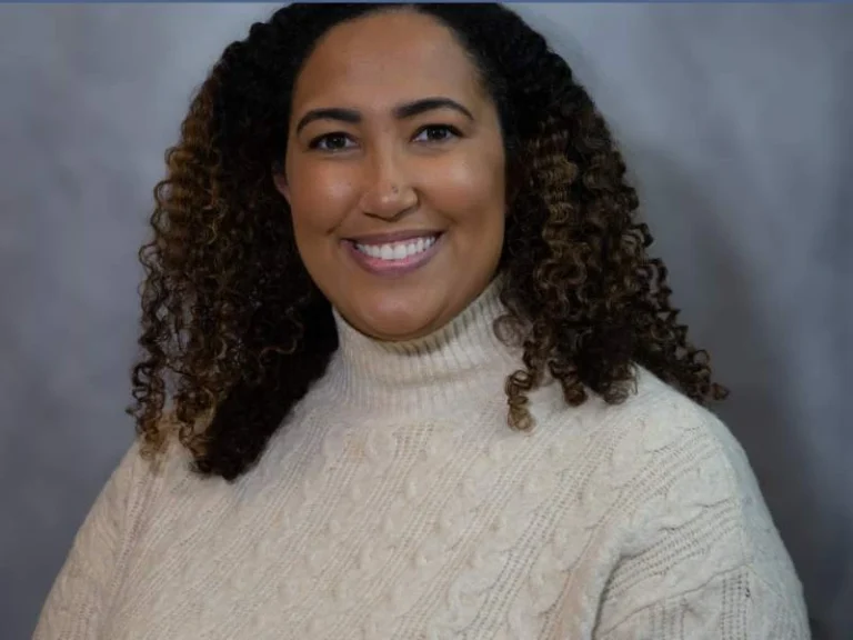 Smiling woman with curly hair wearing a cream cable-knit turtleneck sweater against a gray background.